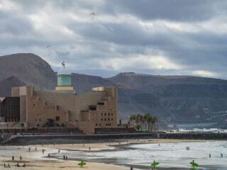 Coastal scene with a tan brick building and green-domed tower by the shore; people on the beach with surfboards and paragliders in the sky.