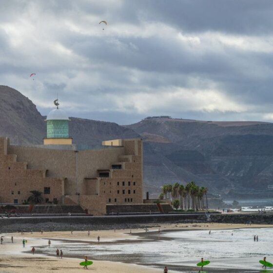 Coastal scene with a tan brick building and green-domed tower by the shore; people on the beach with surfboards and paragliders in the sky.
