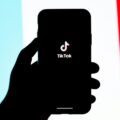 Silhouette of a hand holding a smartphone displaying the TikTok logo against a blue‑white‑red background.