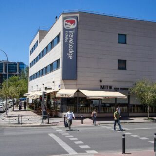 Sunlit street scene featuring a Travelodge hotel with outdoor cafe seating along the sidewalk. Pedestrians walk by on the crosswalks and street corners nearby.