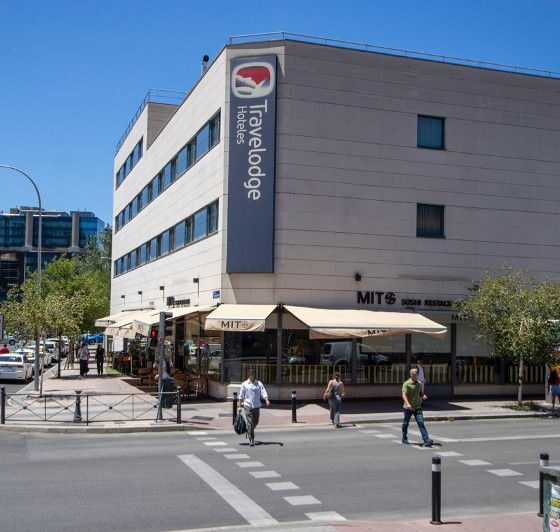 Sunlit street scene featuring a Travelodge hotel with outdoor cafe seating along the sidewalk. Pedestrians walk by on the crosswalks and street corners nearby.