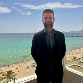 Man with a beard in a dark suit stands on a balcony overlooking a sunny beach and turquoise sea on a clear day.