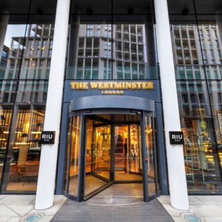 Hotel entrance for The Westminster London, a glass-fronted building with a revolving door and warm lobby light visible inside.