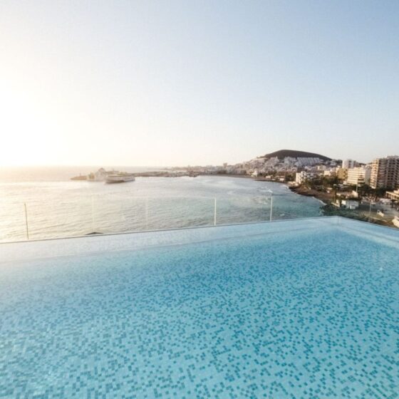 Infinity pool with a mosaic-tile surface overlooking the sea and city lights at sunset, glass railing in the foreground.