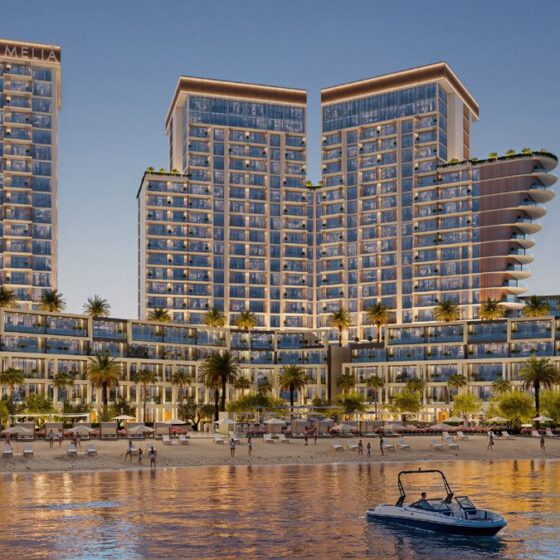 Modern beachfront resort with glass-fronted towers, palm trees, and a sandy beach beside calm water.