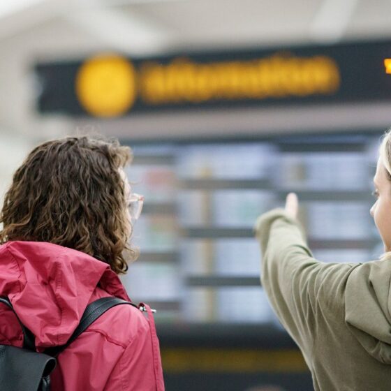 Two travelers—one in a red jacket with a backpack—stand at a station and look at a timetable board, the one on the right pointing ahead