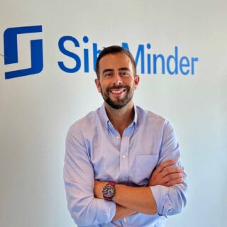 Man in a light blue shirt smiles with arms crossed in front of a wall logo that reads SibMinder or similar brand text in blue.