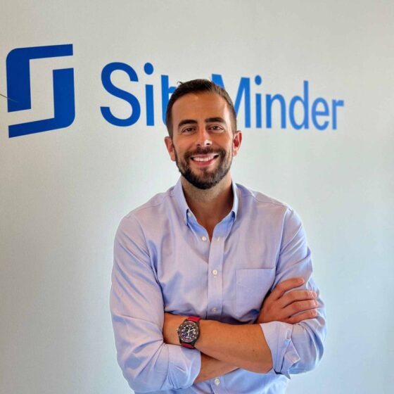 Man in a light blue shirt smiles with arms crossed in front of a wall logo that reads SibMinder or similar brand text in blue.