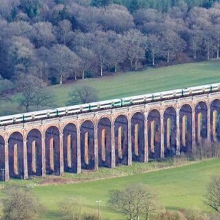 Train crosses a stone arched viaduct over green fields and trees in a rural landscape.