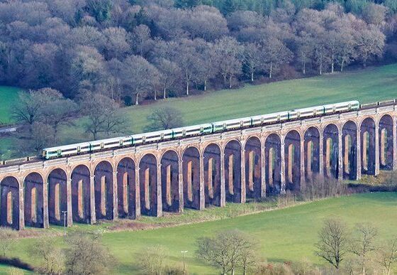 Train crosses a stone arched viaduct over green fields and trees in a rural landscape.