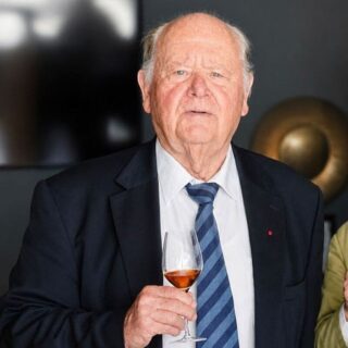Older man in a dark suit and striped blue tie holds a glass of wine at a formal indoor event.