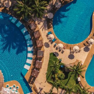 Aerial view of a resort pool complex with curved blue pools, palm trees, and tan lounging decks with umbrellas.