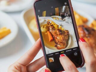 Close-up of hands holding a smartphone, capturing a plated meal with roasted chicken and vegetables at a dining table
