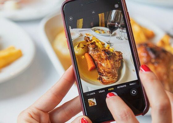 Close-up of hands holding a smartphone, capturing a plated meal with roasted chicken and vegetables at a dining table