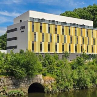 Modern four-story hotel with yellow vertical panels beside a river, surrounded by greenery and a hillside.
