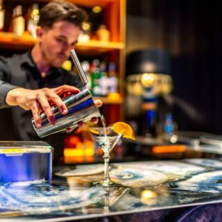 Bartender pours a clear cocktail from a shaker into a martini glass with an orange garnish at a busy bar.