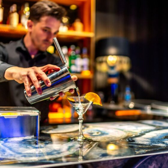 Bartender pours a clear cocktail from a shaker into a martini glass with an orange garnish at a busy bar.