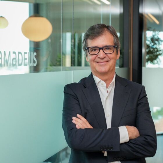 Professional portrait of a middle-aged man in a dark suit with arms crossed, standing in a modern office lobby with glass walls and pendant lights; 'amadeus' logo visible on the glass behind him.