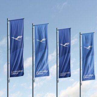 Four blue Lufthansa flags fluttering on poles against a clear sky, each with the white Lufthansa logo and 'Lufthansa Group' text.