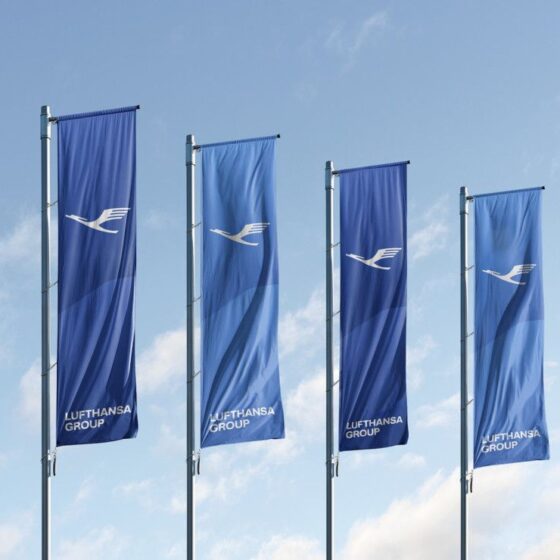 Four blue Lufthansa flags fluttering on poles against a clear sky, each with the white Lufthansa logo and 'Lufthansa Group' text.