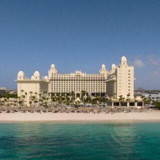 Resort hotel on a sandy beach with turquoise sea, palm trees, and a large beige building complex, RIU sign on a tall nearby tower (beachside vacation scene).