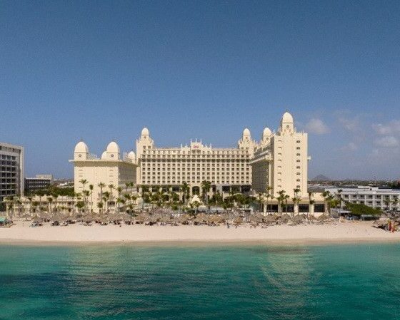 Resort hotel on a sandy beach with turquoise sea, palm trees, and a large beige building complex, RIU sign on a tall nearby tower (beachside vacation scene).