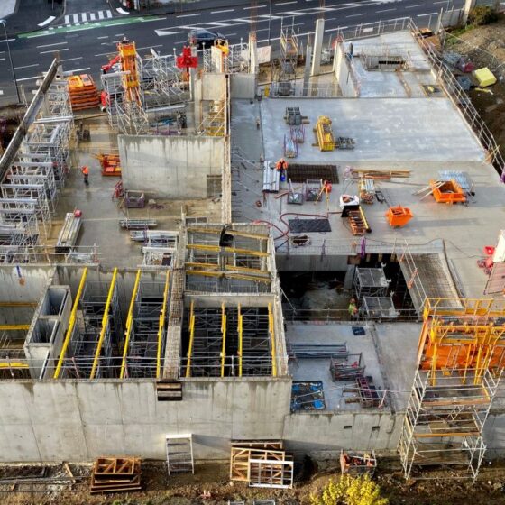 Aerial view of a construction site with unfinished concrete buildings, scaffolding, and yellow safety rails; workers and cranes active amid the site.