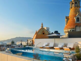 Rooftop pool area with white loungers, umbrellas, and a glass barrier overlooking a city skyline with ornate orange-tiled towers.