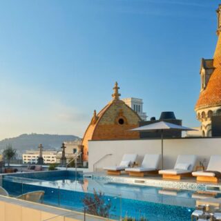 Rooftop pool area with white loungers, umbrellas, and a glass barrier overlooking a city skyline with ornate orange-tiled towers.