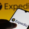 Smartphone in a hand showing the Expedia app logo on a white screen against a bright yellow backdrop.