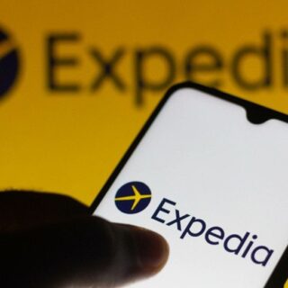Smartphone in a hand showing the Expedia app logo on a white screen against a bright yellow backdrop.