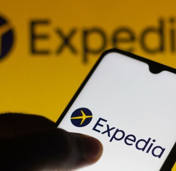 Smartphone in a hand showing the Expedia app logo on a white screen against a bright yellow backdrop.