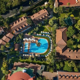 Aerial view of a resort with a central blue pool surrounded by sun umbrellas and palm trees among red-tiled rooftops and green landscaping.