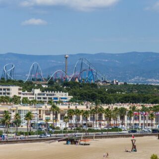 Coastal city scene with a sandy beach in the foreground, palm-lined promenade, and a distant roller coaster set against hills and blue sky.