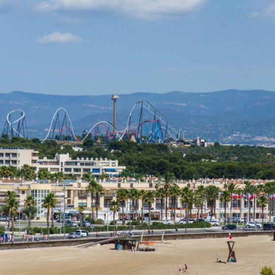 Coastal city scene with a sandy beach in the foreground, palm-lined promenade, and a distant roller coaster set against hills and blue sky.
