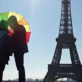 Two people stand behind a large multicolored rainbow umbrella silhouetted against the Eiffel Tower and Paris skyline in daylight.