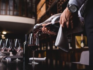 A waiter pours red wine from a bottle into a glass at a bar.