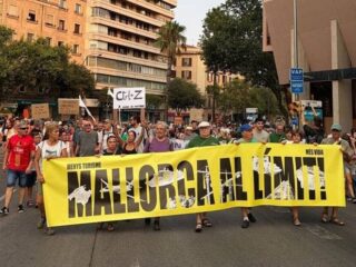 Group of protesters marching down a city street holding a large yellow banner that reads 'Mallorca al dimite!'