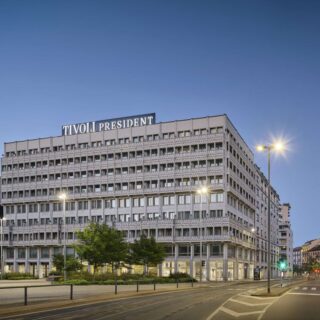 Exterior of the Tivoli President building, a mid‑rise concrete office block with a grid of windows, at blue hour by a street with lamps and tram tracks