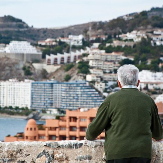 Older man with gray hair in a green jacket stands at a stone railing, gazing over a coastal town built on hills with white and terracotta buildings by the sea.