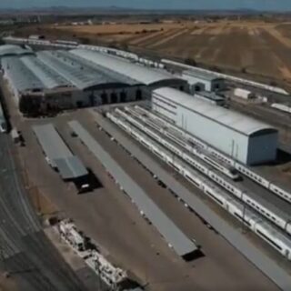 Aerial view of a large rail yard with multiple tracks, long passenger trains, and white warehouse buildings in a dry, agricultural landscape nearby.