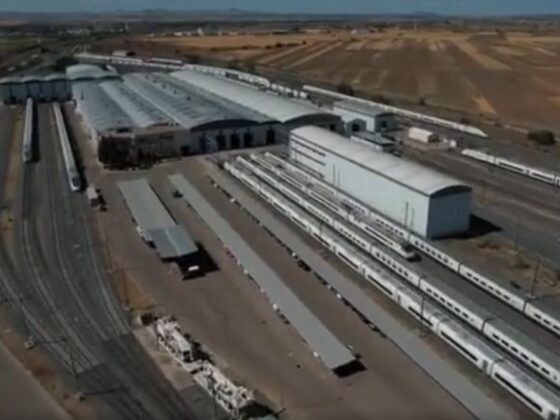 Aerial view of a large rail yard with multiple tracks, long passenger trains, and white warehouse buildings in a dry, agricultural landscape nearby.