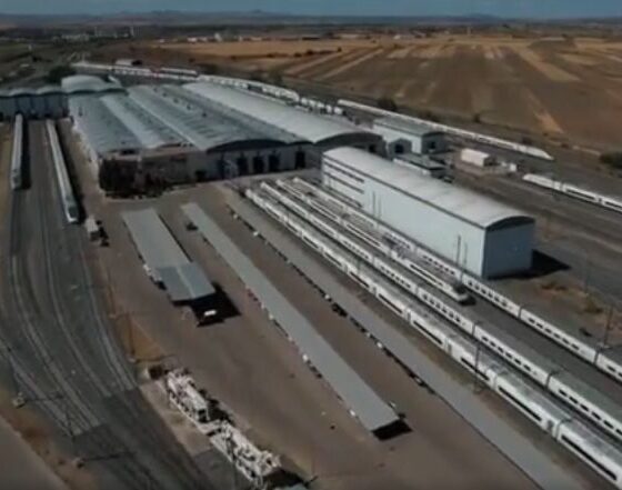 Aerial view of a large rail yard with multiple tracks, long passenger trains, and white warehouse buildings in a dry, agricultural landscape nearby.