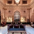 Luxurious hotel lobby with marble walls, arched doorways, and dark leather chairs around a central blue ottoman.