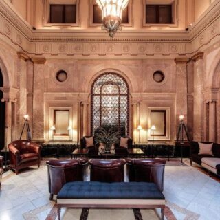 Luxurious hotel lobby with marble walls, arched doorways, and dark leather chairs around a central blue ottoman.