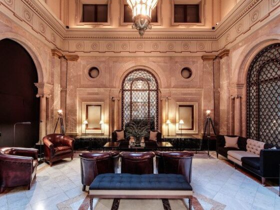 Luxurious hotel lobby with marble walls, arched doorways, and dark leather chairs around a central blue ottoman.