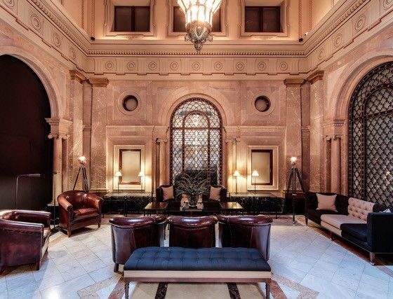Luxurious hotel lobby with marble walls, arched doorways, and dark leather chairs around a central blue ottoman.