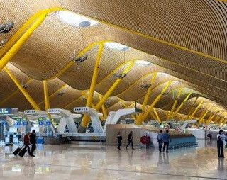 Interior of an airport terminal with yellow curved beams, a glossy floor, and travelers with luggage near check-in desks.