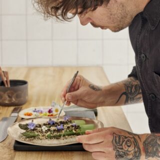 Tattooed chef carefully plates a dish with asparagus and edible flowers on a wooden counter at the kitchen station.