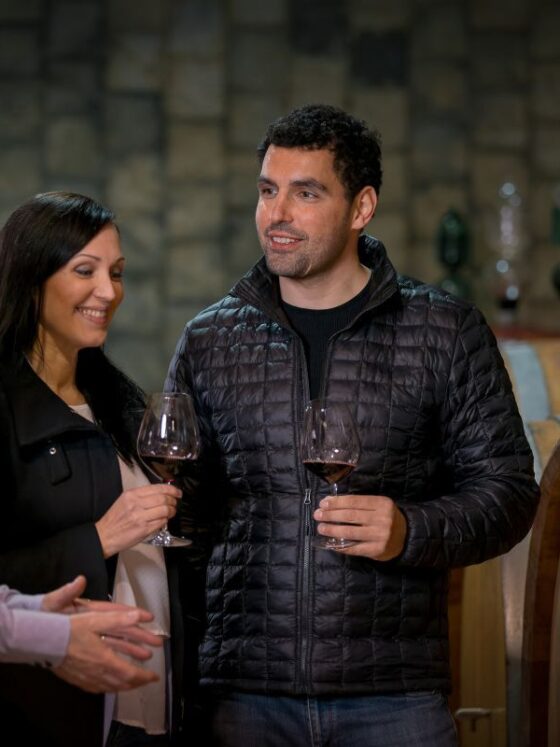 Three people sample wine in a cellar, holding glasses near wooden wine barrels and enjoying conversation.
